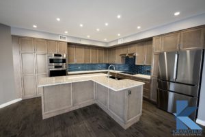 Modern kitchen with light wood cabinets, stainless steel appliances, a large island with a sink, and blue tile backsplash. The floor is dark wood, and recessed lighting is overhead.